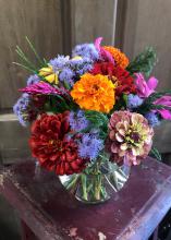 Orange, red, pink, purple, and yellow flowers are arranged in a glass bowl that sits atop a small, red table.