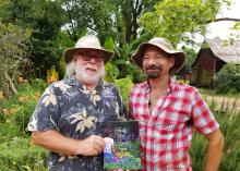 Two men in hats stand in a garden setting and hold a book on display.