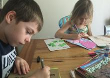 A boy and girl sit at a wooden table and use markers to draw on different colored sheets of paper.
