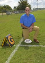 A man kneels on a football practice field beside a football.