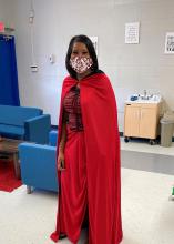 A woman in a red cape and mask stands in a classroom.