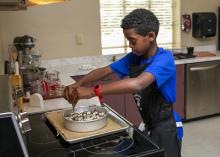 A boy standing at a stove drizzles chocolate over a dessert.