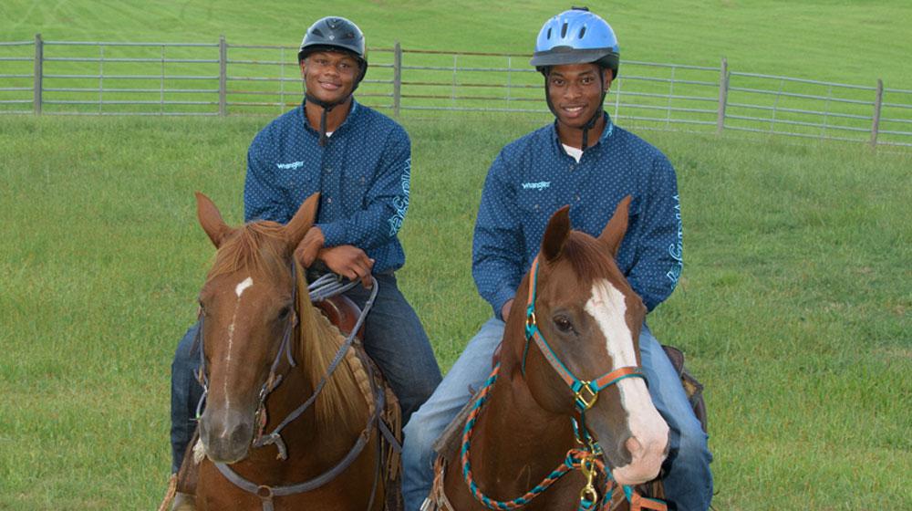 Two boys sit on two horses facing the camera. Both boys are wearing blue shirts and safety helmets.