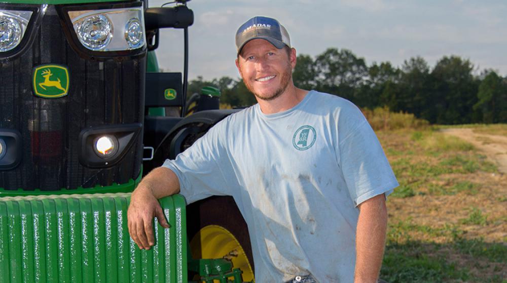 A man wearing a blue t-shirt and blue jeans leans against a green tractor.