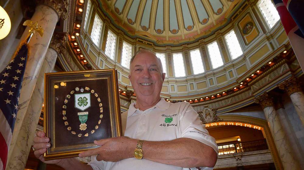 A man with a white 4-H shirt and khaki pants stands in the Mississippi capitol building holding a golden frame with the 4-H green four-leaf clover in the center. Surrounding the logo in a circle are more than 20 4-H pins. 
