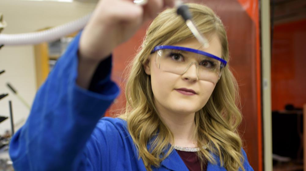 A blonde young woman wearing a blue lab coat and safety glasses holds a glass dropper up to the camera.