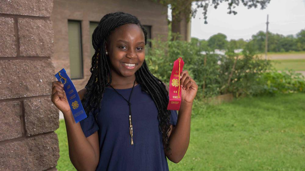 4-H’er Ashantis Wigley Holding Award Ribbons A teen girl wearing a blue shirt stands next to the corner of a brick wall and in front of green bushes. She holds a blue ribbon in her right hand and a red ribbon in her left.