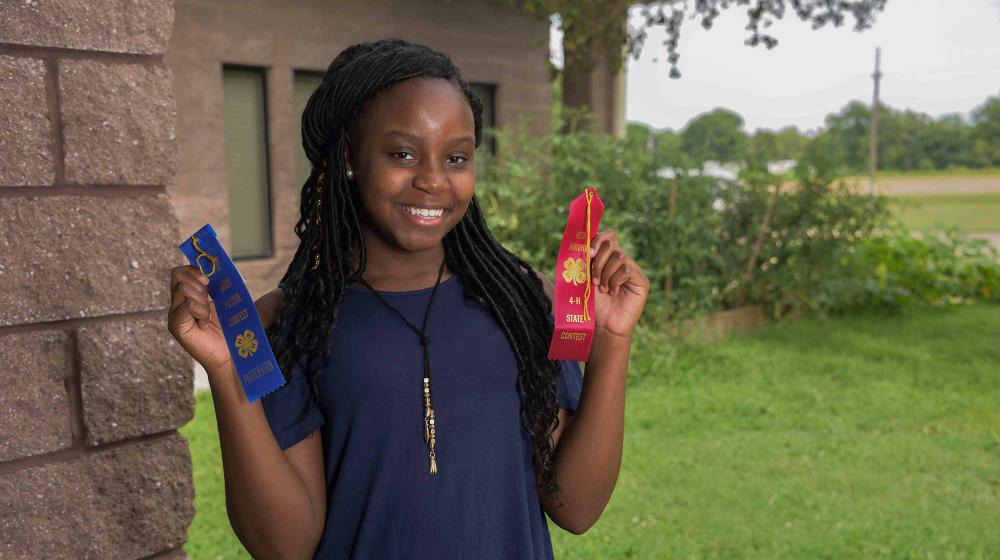 A teen girl wearing a blue shirt stands next to the corner of a brick wall and in front of green bushes. She holds a blue ribbon in her right hand and a red ribbon in her left. 