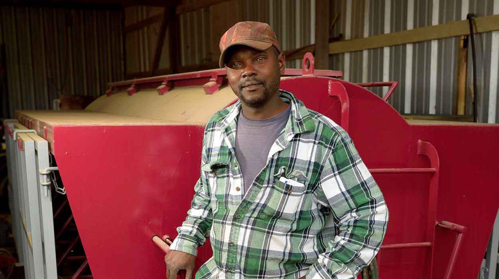 A man wearing a green and white plaid shirt, blue jeans, and a brown baseball cap stands in front of a bright red machine parked inside a metal building.  