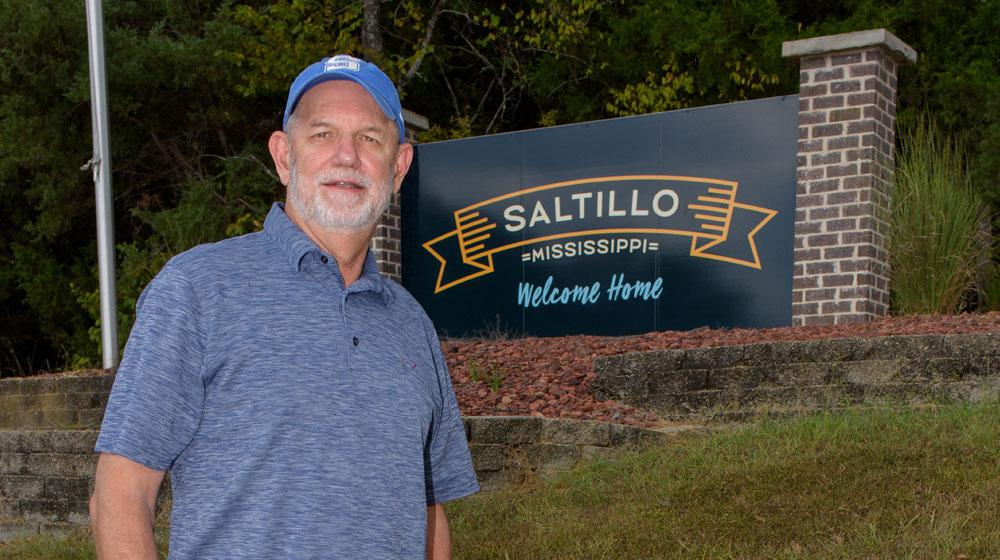 A man stands in front of a sign that reads, “Saltillo Mississippi, Welcome Home.”