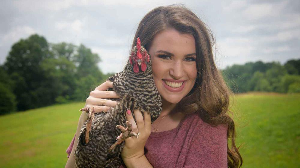 A young woman with brown hair and a pink shirt stands smiling while holding a black and white speckled chicken next to her face. 