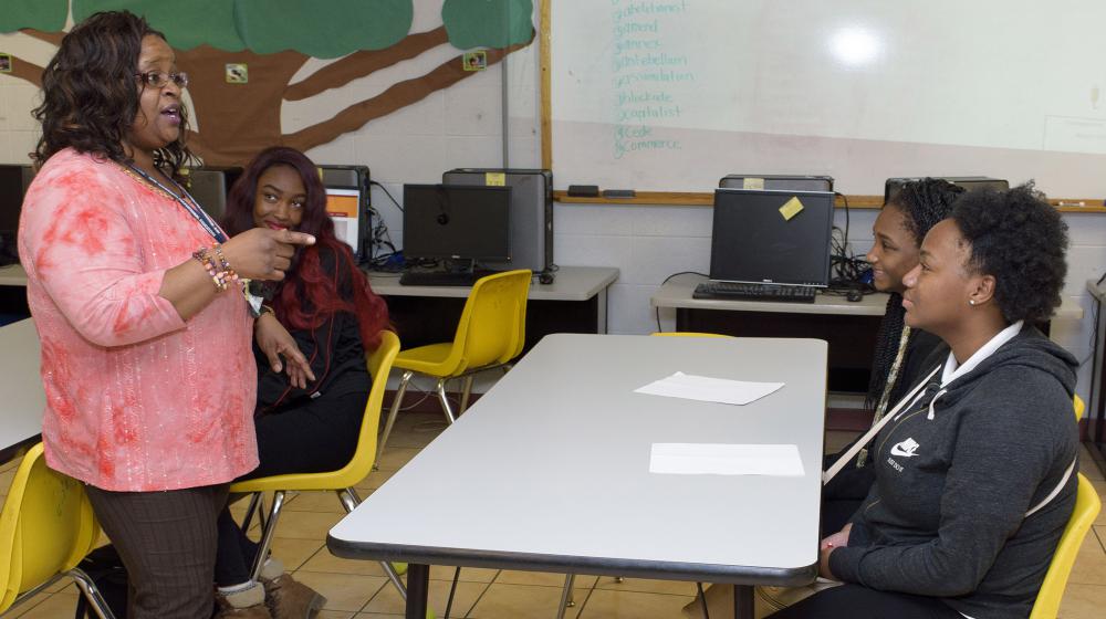 A woman, leftmost, wearing a coral blouse and brown slacks, points and talks to two teens sitting in yellow chairs at a gray table.