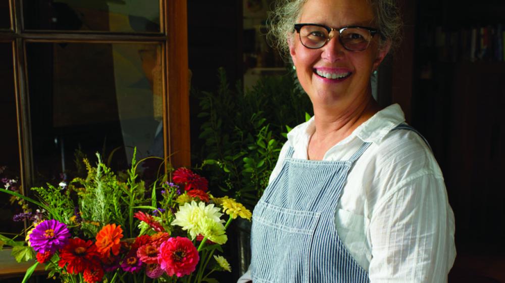 Woman with glasses stands smiling beside a flower arrangement