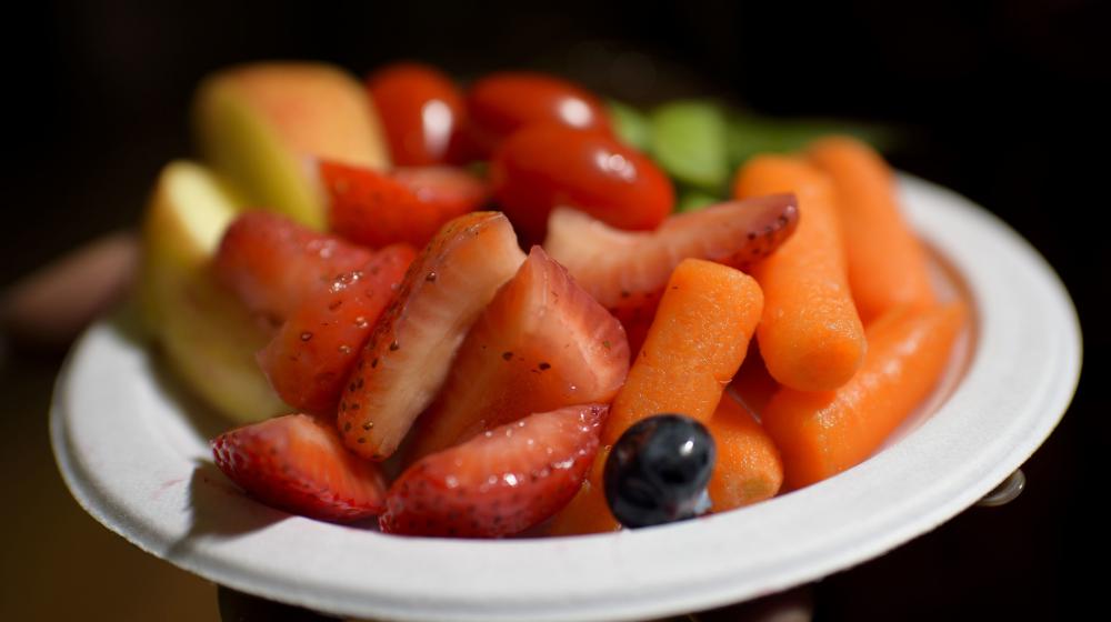 A white bowl contains red grape tomatoes, sliced strawberries, sliced red apples, green sugar snap peas, orange carrots, and a dark purple grape.