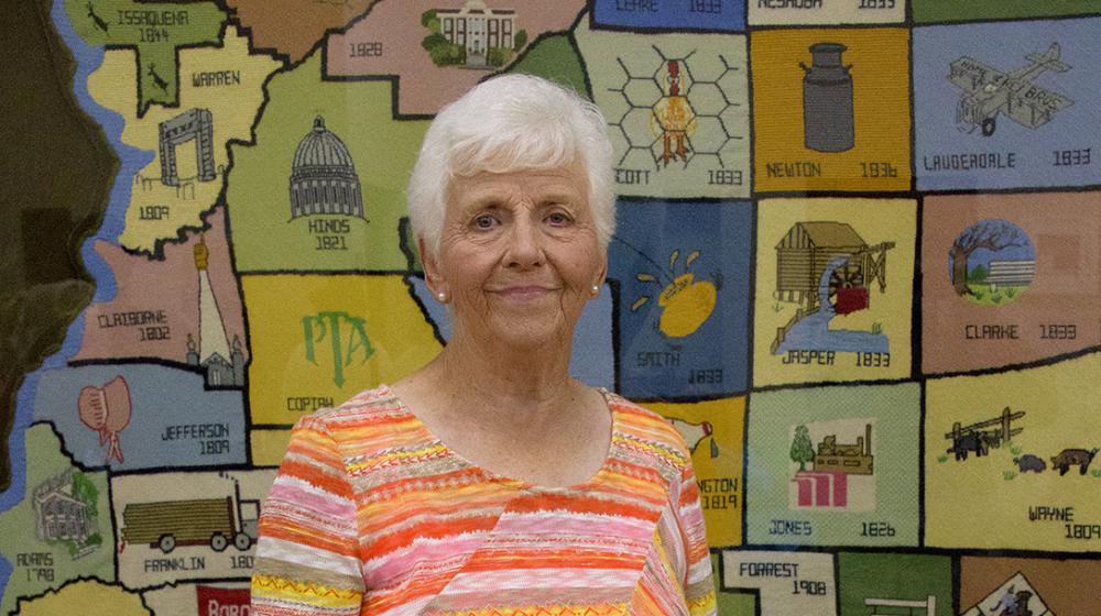 Rae Clarke, Mississippi Homemaker Volunteer An elderly woman wearing an orange striped shirt stands in front of a large, multicolored, needlepoint county map of Mississippi.
