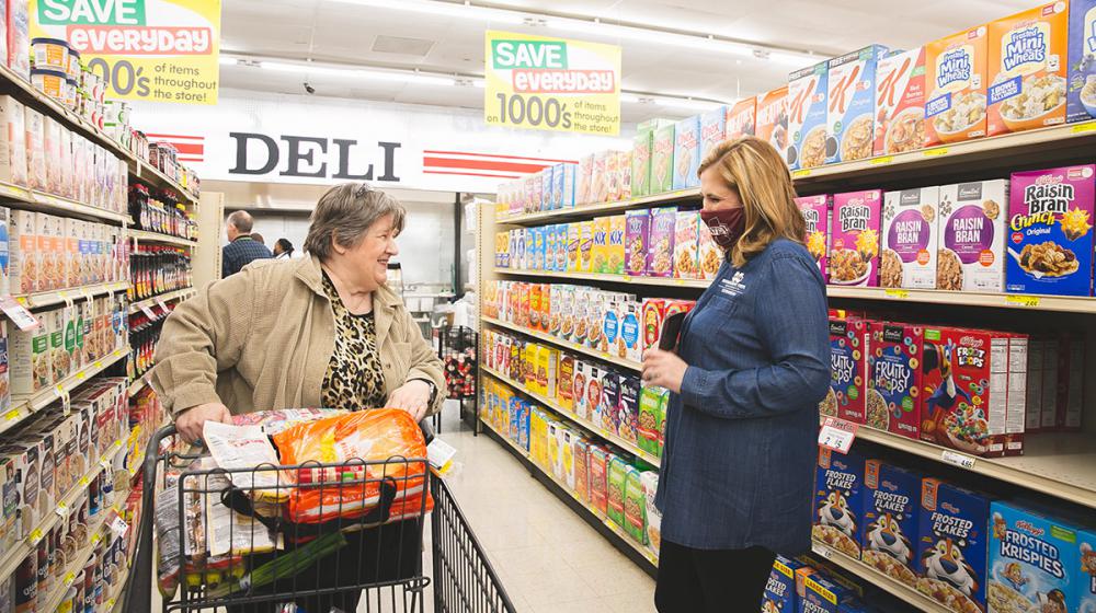 A woman wearing a Mississippi State University mask happily speaks with a woman pushing a grocery cart down an aisle of cereal in a grocery store.