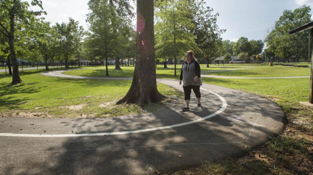 A woman walking on a sidewalk curved around a tall tree.