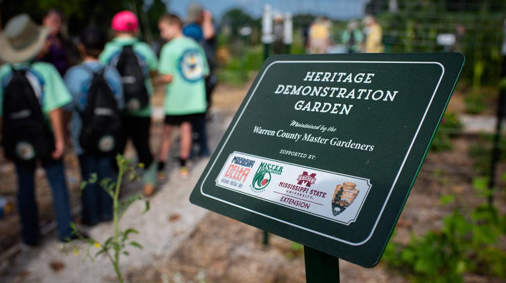 A sign marking the entrance to the Heritage Demonstration Garden with children standing in the background.