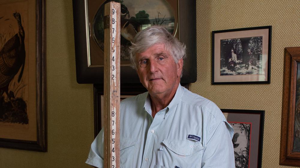 A man holding a large wooden measuring stick stands in front of a wall with several framed photos.