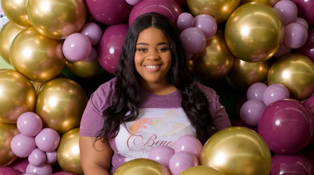 A smiling young woman wearing a shirt that reads “Beyond the Arch” stands next to several balloons and jars of party supplies.
