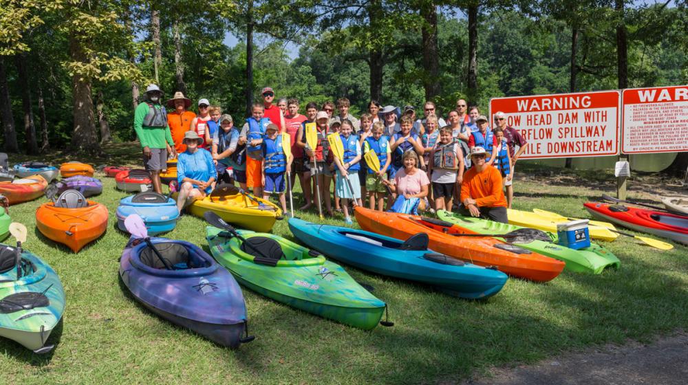 A large group photo of several young children and adults wearing kayaking gear and several kayaks of different colors in the grass in front of them.