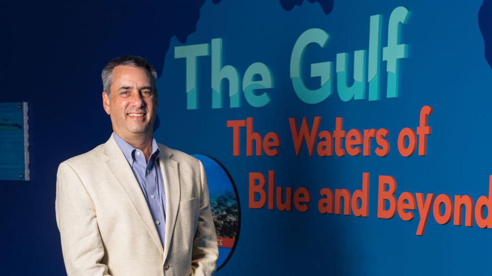 A man wearing dress clothes stands in front of a blue wall and smiles with his hands clasped in front of his body.