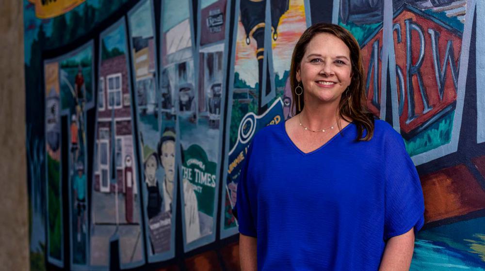 A smiling woman in a blue shirt stands in front of a wall mural.