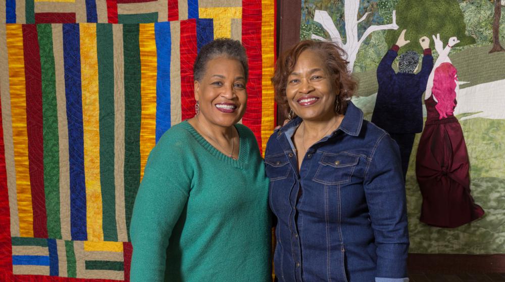 Two Black women smiling and standing in front of a hanging multicolored quilt.