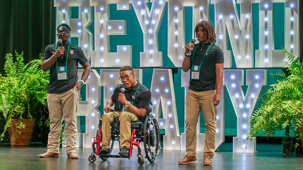 Three teens sing on stage, with the central performer seated in a wheelchair. Large, illuminated letters, arranged to form the words “Beyond Ready,” are in the background.