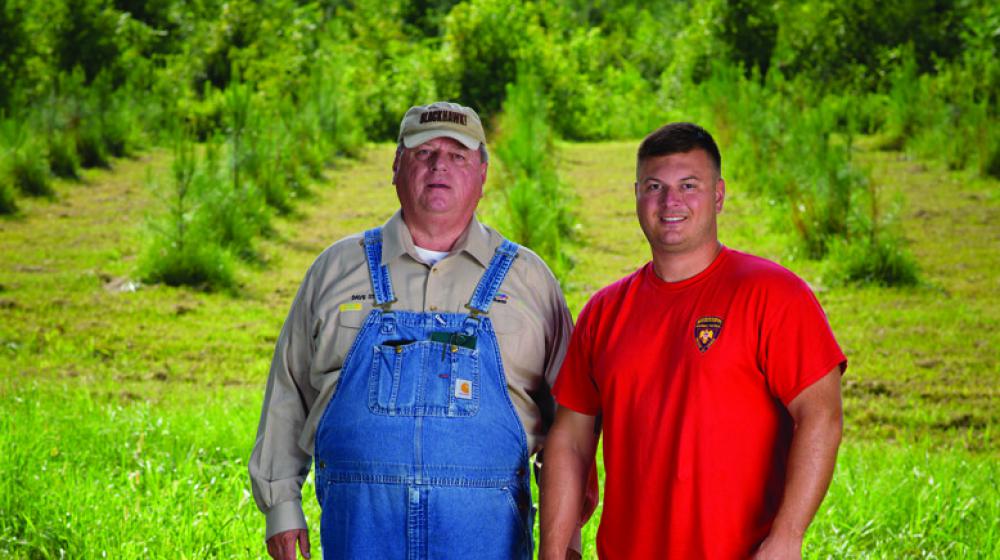 • A man in overalls stands beside a man in a red T-shirt in front of a tree farm