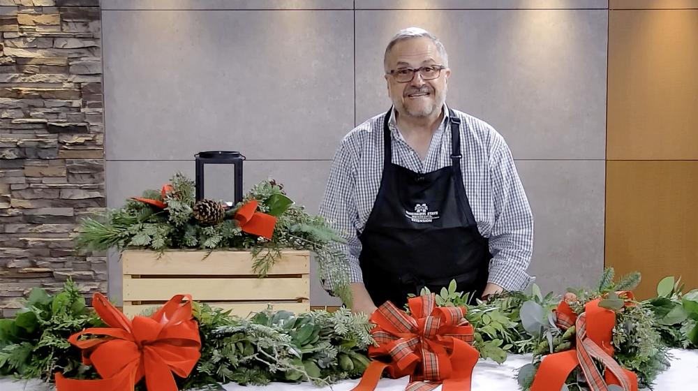 A man, smiling, standing in front of a candle centerpiece and behind a table with bows, evergreen door swag and evergreen garland.