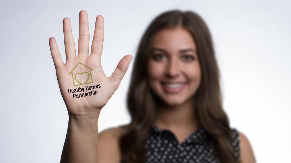 A young woman with dark hair extends her palm forward with the Healthy Homes Initiative logo on her palm.