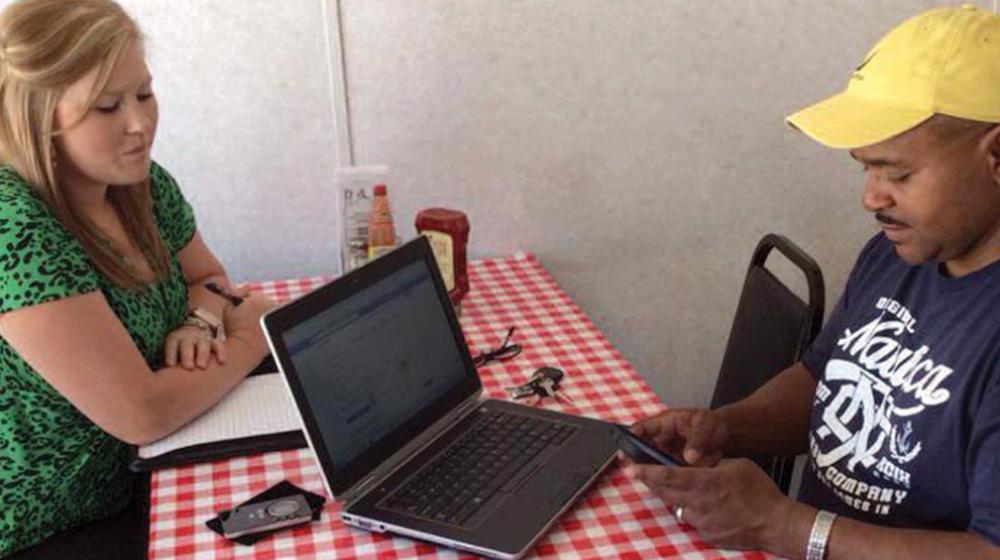 A woman sits on one side of a table, and a man with a laptop sits opposite from her.