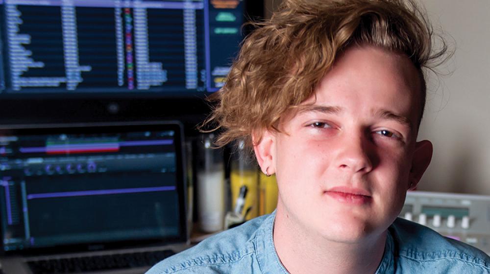 A young man sitting at a desk with computer monitors behind him.