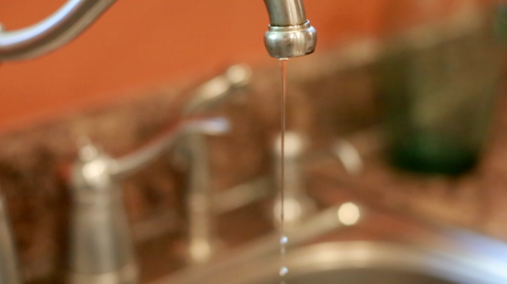 A thin stream of water drips from a stainless steel kitchen sink faucet.