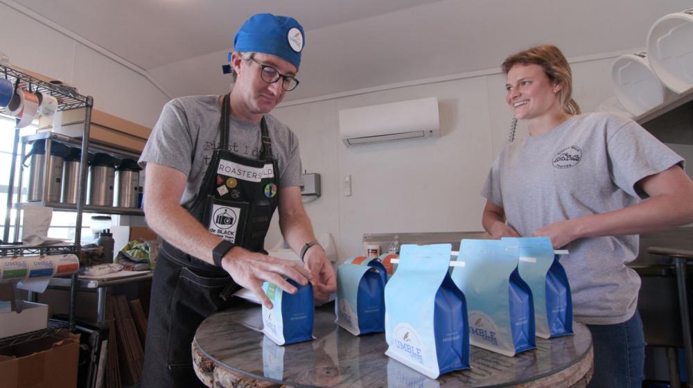 : A man with a blue cap and glasses grabbing a blue bag of coffee for a blonde, smiling woman.
