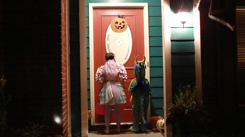Two children waiting at a well-lit door for Halloween treats.