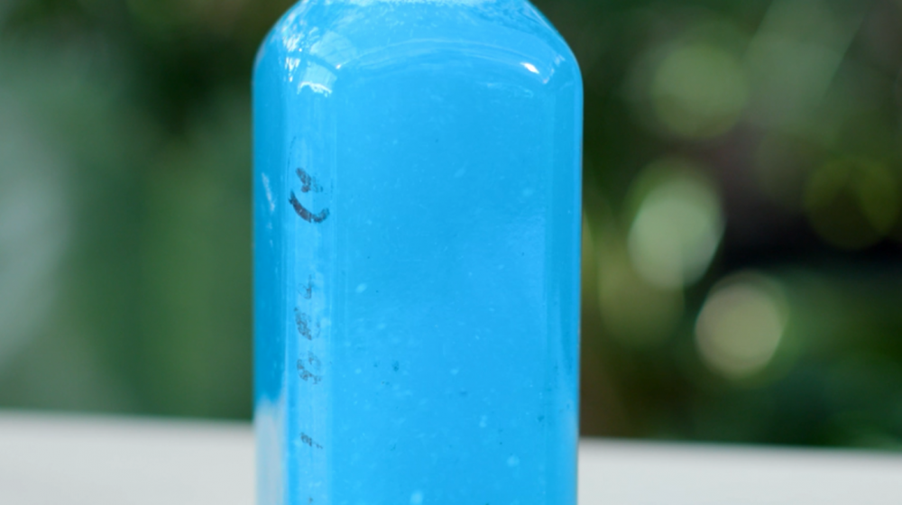 Close-up of a portion of a clear bottle filled with bright blue liquid that is water-soluble fertilizer dissolved in water, against a blurred dark green background.