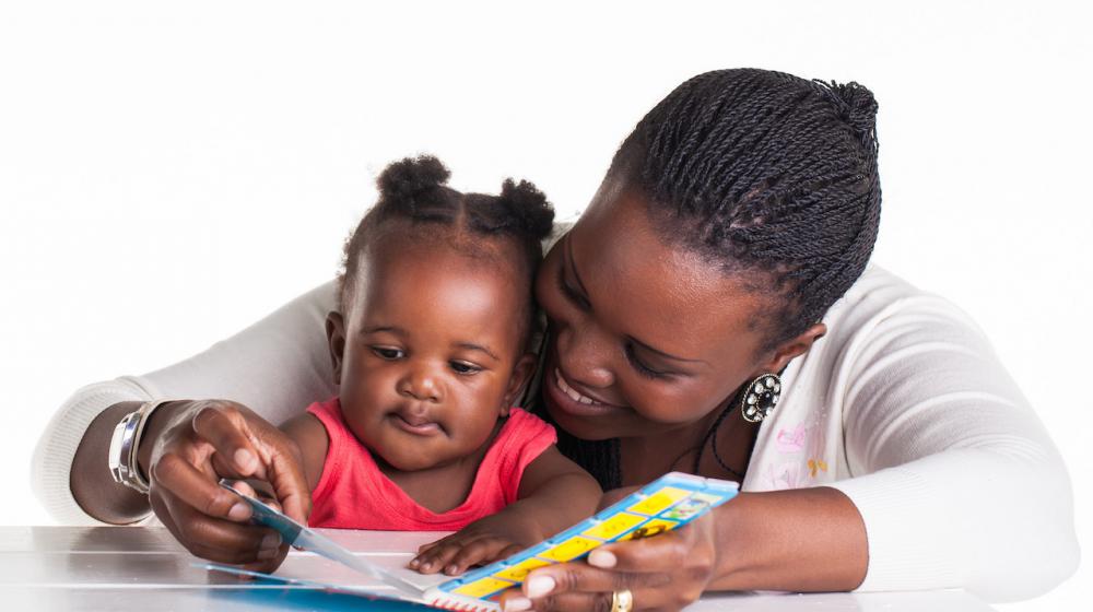 An African American woman with braided hair and wearing a white sweater shares a board book with her infant daughter who wears a coral shirt.