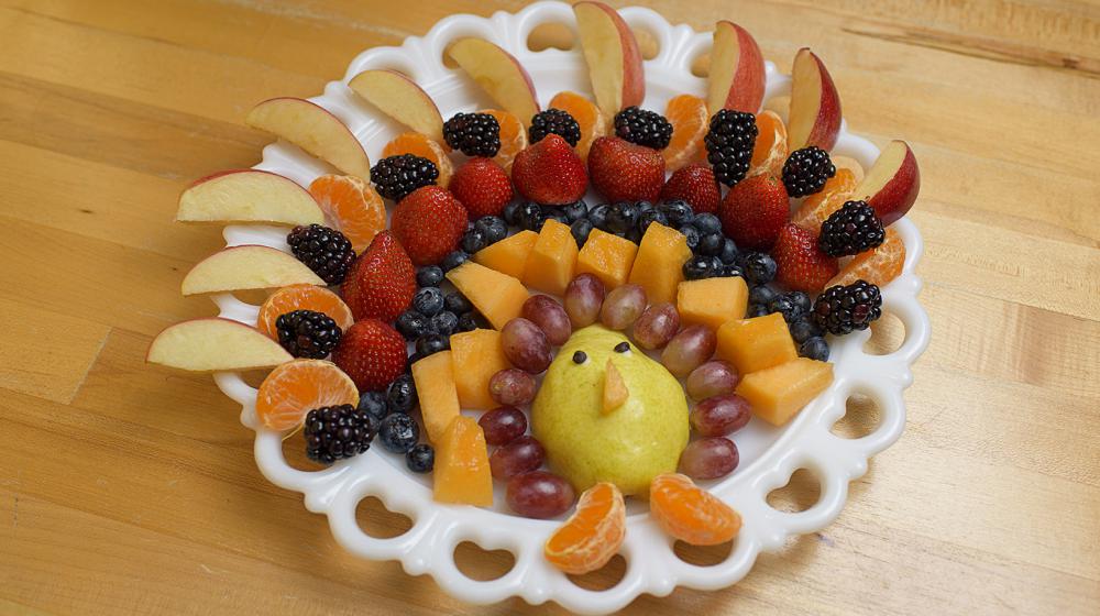 Several rows of fruit including apple slices, tangerine segments, blackberries, blueberries, cantaloupe, strawberries, grapes and a pear are arranged on a white platter in the shape of a turkey.