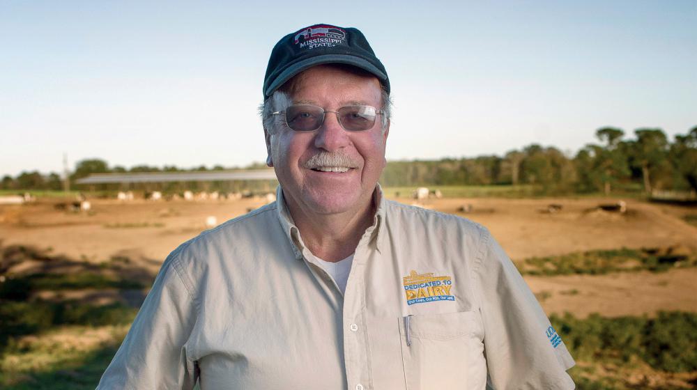 A smiling man with a "dedicated to dairy" logo on his shirt, stands in a pasture.