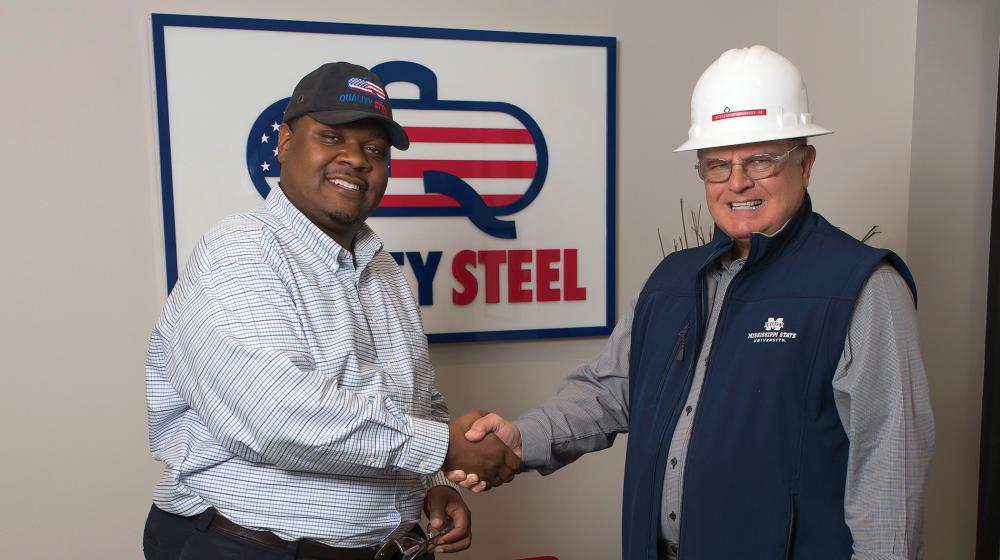 Two men shake hands in front of a Quality Steel sign.