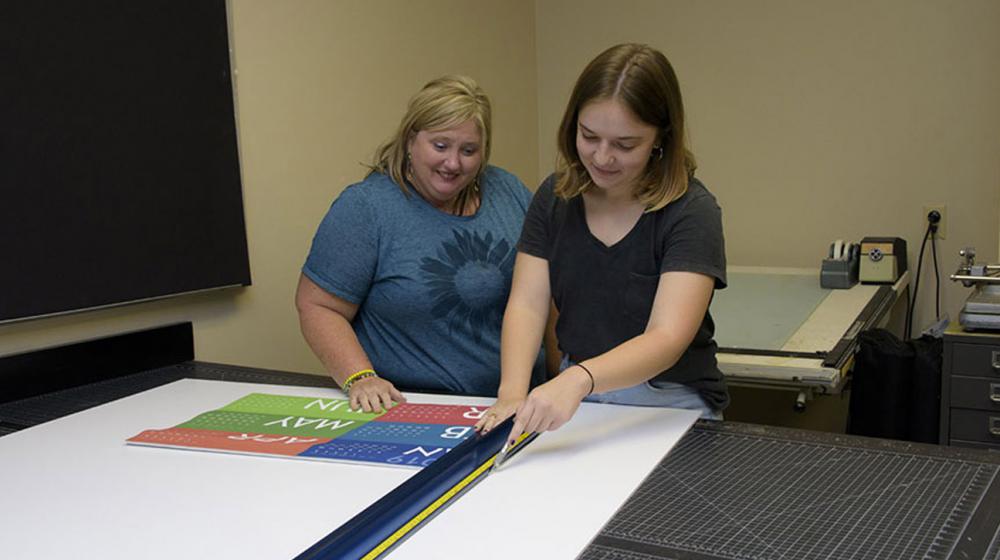 A woman with blonde hair and a blue shirt smiles as a younger brunette lady wearing a black shirt and measuring materials on a board.