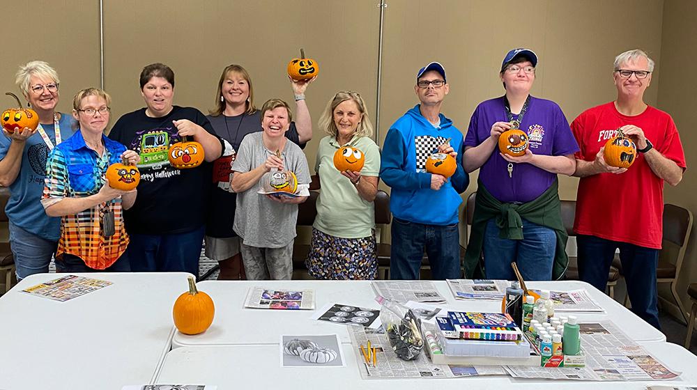 Nine people standing behind a crafts table holding pumpkins with pained faces.