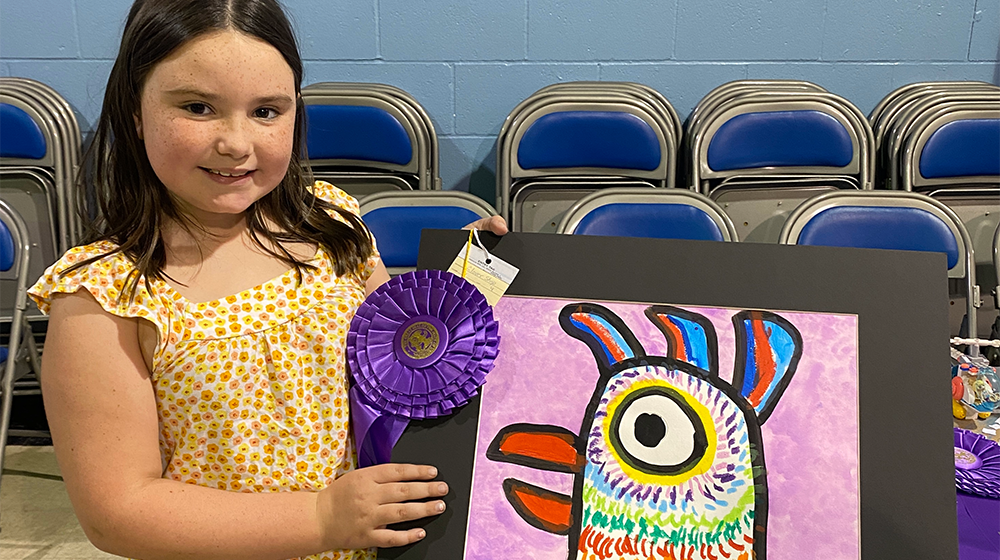 Young girl showing a blue ribbon winning painting of a rooster.