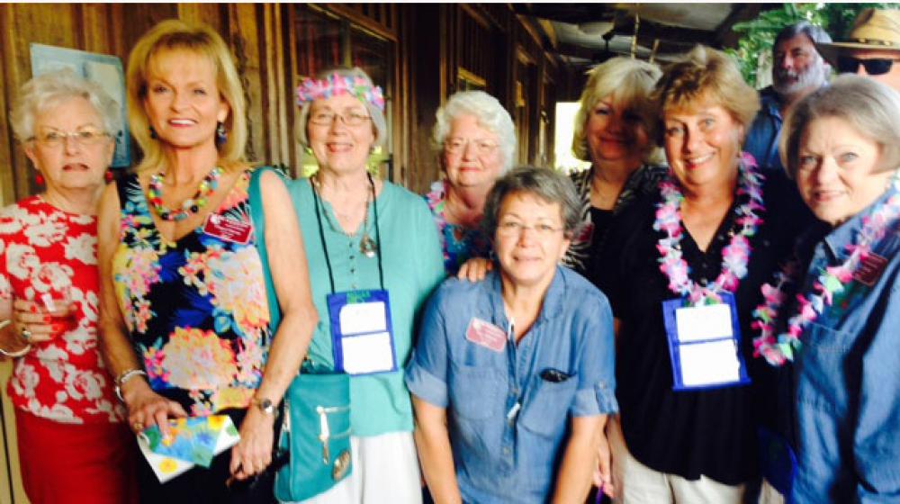 Eight older women wearing Hawaiin themed clothes stand next to each other smiling.