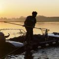 man standing in a motorboat using a large fishing net to catch catfish against an orange sunrise 