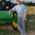 A man wearing a blue t-shirt and blue jeans leans against a green tractor.