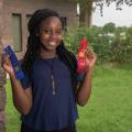 A teen girl wearing a blue shirt stands next to the corner of a brick wall and in front of green bushes. She holds a blue ribbon in her right hand and a red ribbon in her left. 