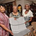 : On the left, a younger woman wearing a patterned pink shirt holds one side of a crocheted grey shirt shirt. In the middle, and older woman smiles. On the right, another older woman with grey hair and a crocheted piece of clothing smiles and holds the shirt. 
